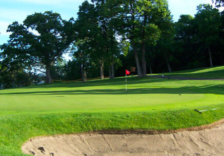 A lush golf course with a sand bunker in the foreground, a red flag on the putting green, and tall trees in the background beneath a clear blue sky.