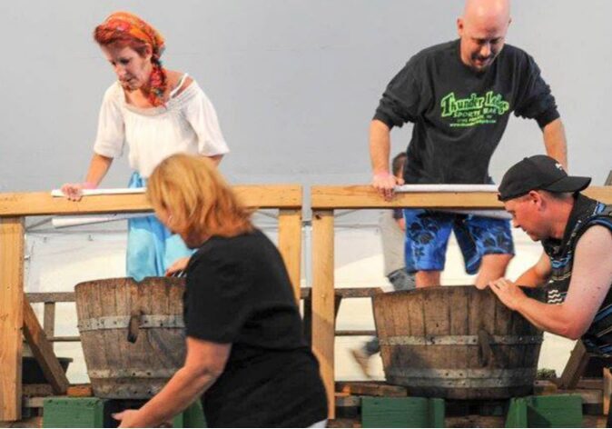 Two men and a woman grapple grape stomping in wooden barrels on a raised platform at an Alexandria event. Another woman stands in front, slightly blurred, as they enjoy one of the area’s favorite autumn activities.