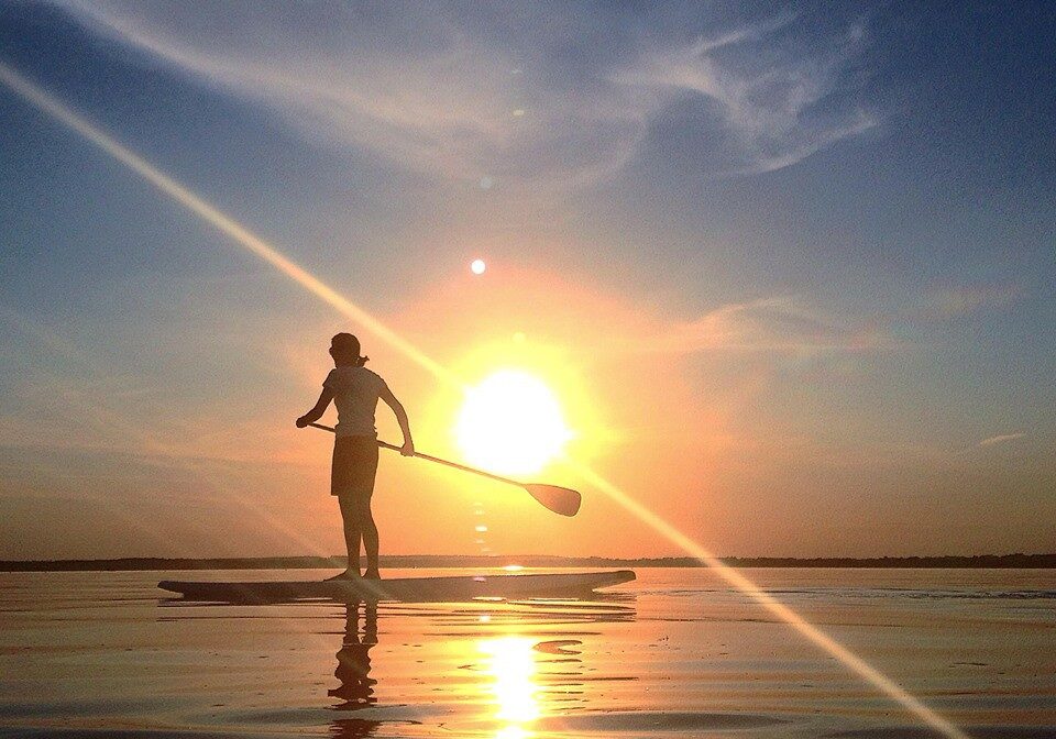 A person stands paddling on a board over calm water during sunset, with the sun low in the sky, casting a golden reflection—a serene Hang Loose MN moment—on the water and soft clouds in the background.