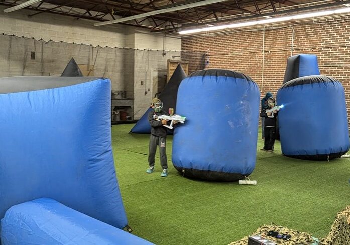 Three children wearing goggles play with toy guns in an indoor arena called Hildebrandt Hideaway, dodging behind large blue inflatable barriers and running across artificial turf, surrounded by brick and concrete walls.