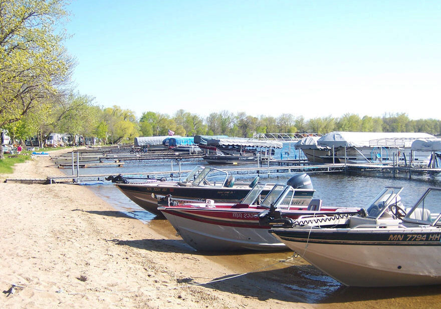 Several motorboats are lined up along a sandy lakeshore near docks at Head of the Lakes Resort Campground. Trees with green leaves border the lake, and covered boat slips are visible in the background under a clear sky.