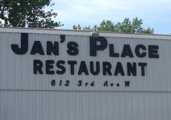The sign for Jan’s Place Restaurant displays bold black letters on a light-colored building, with “612 3rd Ave W” written below Jan’s Place. Trees and blue sky can be seen in the background.
