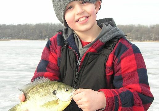 A smiling child in a gray beanie and red plaid jacket holds a fish while standing on a frozen lake, with bare trees in the background—another memorable catch with JS Guide Services.