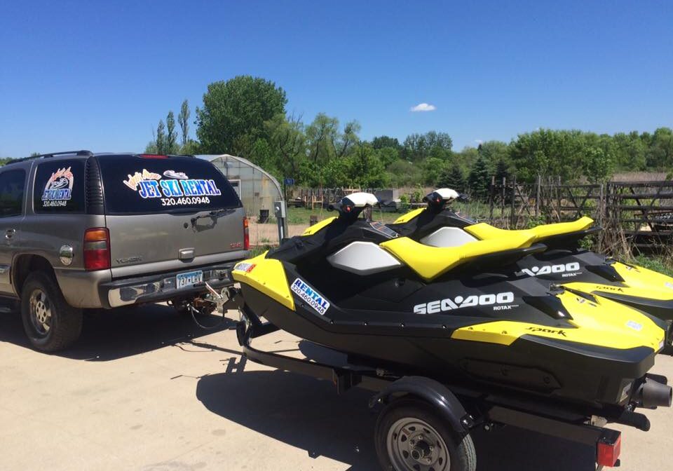 A black SUV with jet ski rental signage is towing a trailer loaded with two yellow and black Sea-Doo jet skis on a sunny day. Trees and a clear blue sky form the backdrop.