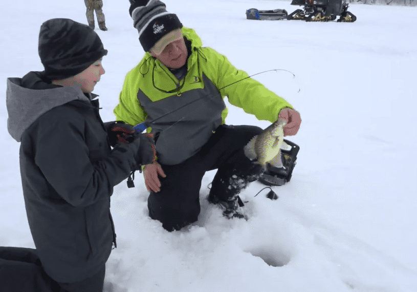 A man kneels on snow by an ice fishing hole, proudly holding up his catch, while a child in winter clothing stands beside him. Fish, snowmobiles, and gear dot the snowy landscape in the background.