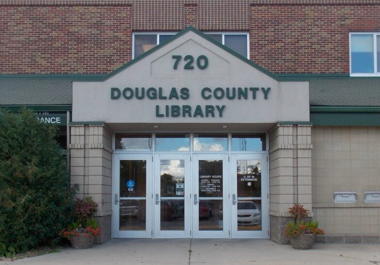 Exterior view of the Douglas County Library, featuring glass entrance doors, a “Douglas County Library” sign above street number 720, surrounded by shrubs and a classic brick building facade.