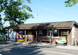 A small rustic wooden building labeled "Office" sits by a lakeside road at Linwood Resort, with a candy machine, an ice machine, and a children's ride out front. Trees and water provide a scenic backdrop to this charming campground spot.