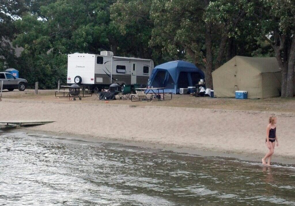 A woman in a black swimsuit walks along the shoreline at Lucky Acres Campground; behind her are tents, a camper trailer, picnic tables, and trees at this scenic lakeside campsite.