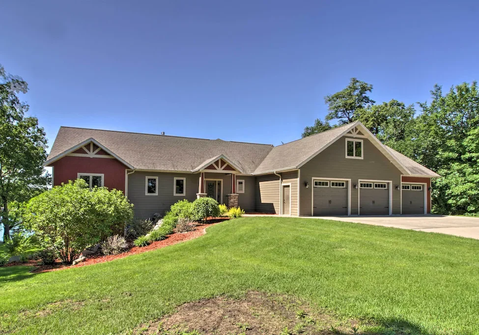 Luxury on Lake Latoka awaits in this single-story house with gray siding, a three-car garage, and a gabled roof, all nestled amid green lawns, landscaping, and trees under a clear blue sky.