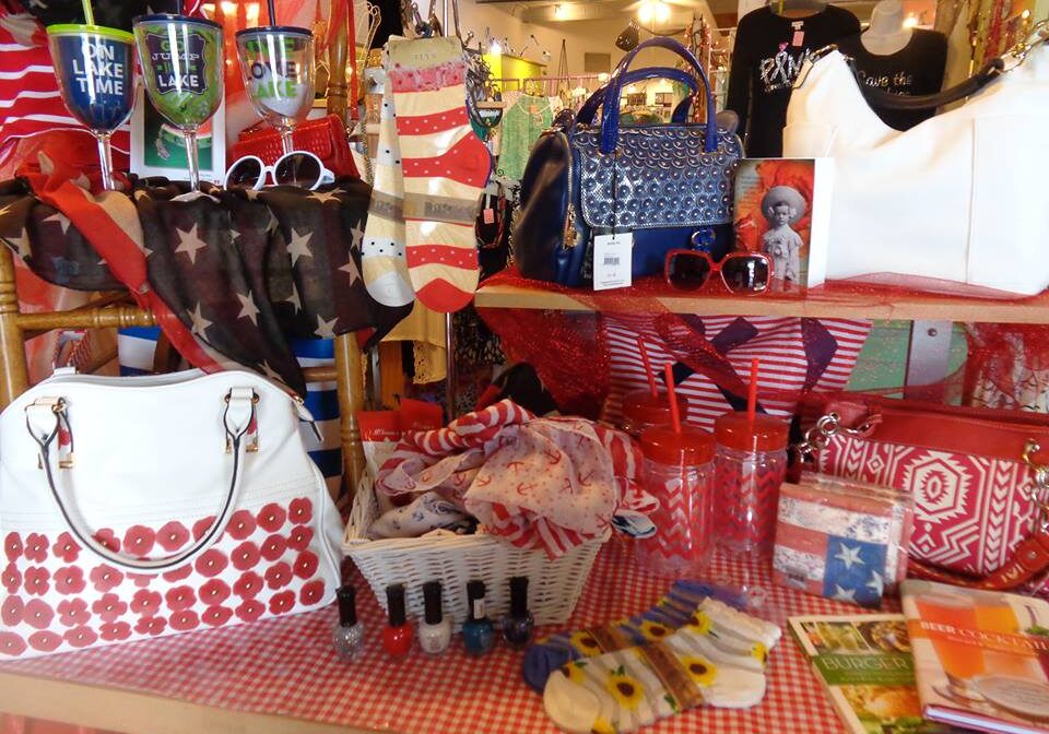 A magpie-worthy display of patriotic-themed gifts—handbags, socks, scarves, sunglasses, tumblers, nail polish, and books—mostly in red, white, and blue hues, arranged on a red checkered tablecloth in the store.
