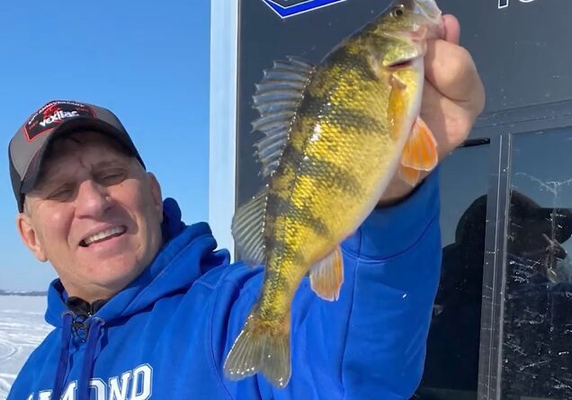 Mike Frisch, wearing a blue hoodie and cap, holds up a yellow perch fish and smiles while standing outside on a snowy, frozen lake.