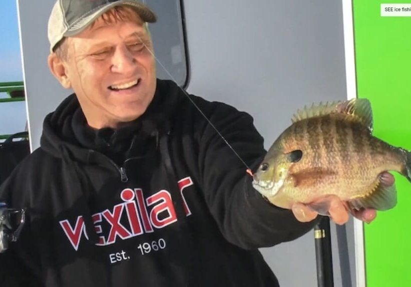 A smiling man, mike frisch, wearing a black "Vexilar" hoodie and cap, holds up a freshly caught fish on a fishing line, standing in front of a white and green background.