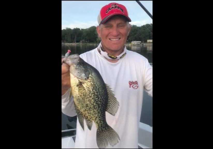A man in a white long-sleeve shirt and red cap smiles while holding up a large speckled fish on a boat, with water and trees in the background—capturing a classic Mike Frisch fishing moment.