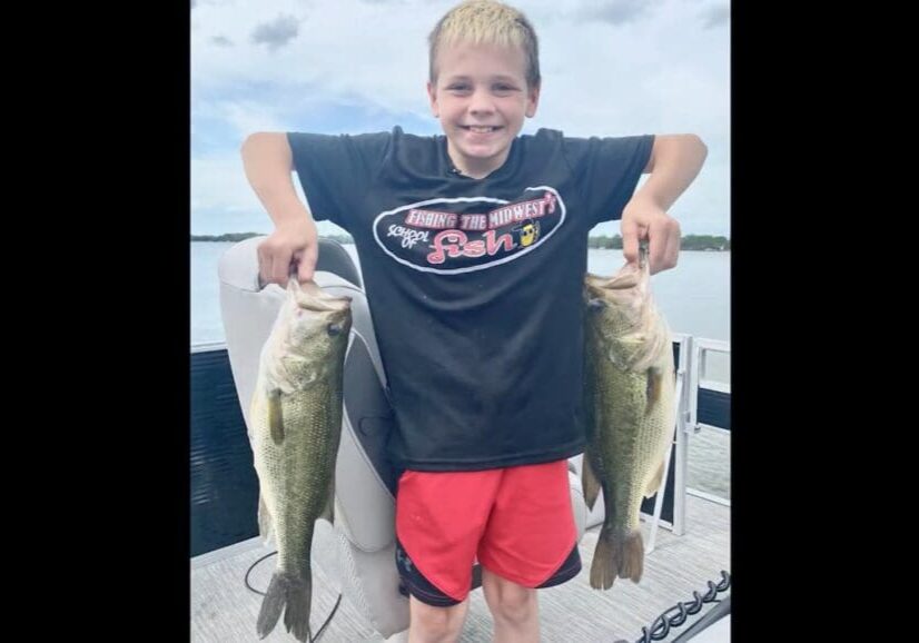 A smiling boy stands on a boat, proudly holding up two large fish—one in each hand—after a successful day of fishing. He wears a black T-shirt and red shorts, with water and sky stretching out behind him.