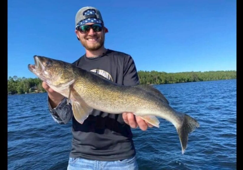 A smiling man, Mike Frisch, wearing sunglasses and a cap, holds a large fish on a boat, with blue water and a forested shoreline in the background under a clear blue sky.