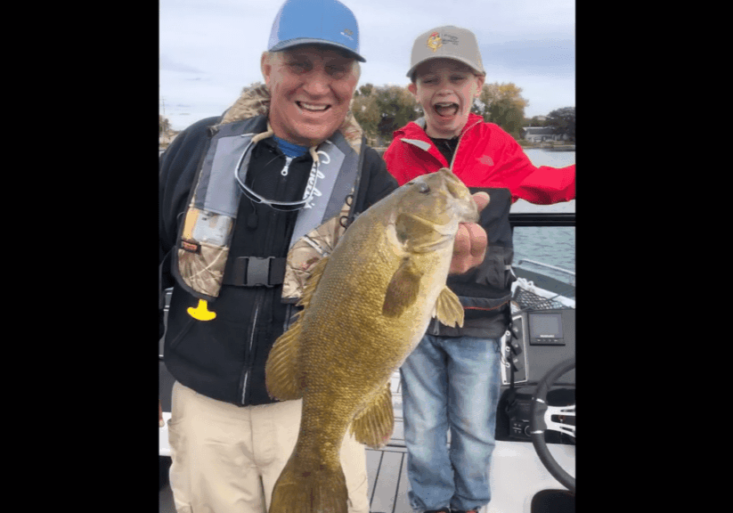 An older man and a young boy smile excitedly on a boat, proudly holding up a large fish they caught while fishing. The boy wears a red jacket and cap, the man sports a blue cap and life vest, with water and trees in the background.
