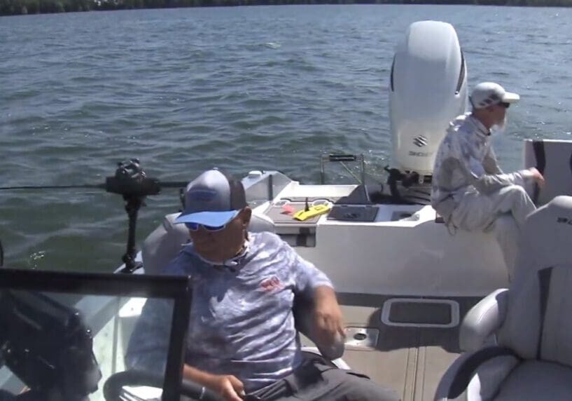 Two people in fishing attire sit on a small motorboat on a lake. One, possibly following Mike Frisch’s tips, is at the back near fishing gear while the other sits near the front, both under a sunny sky with calm water.