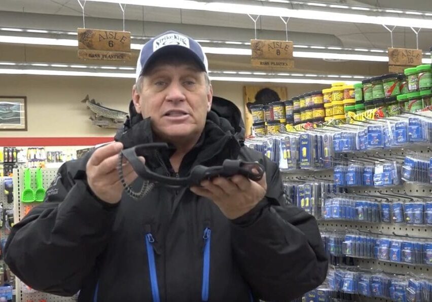 A man in a cap and jacket stands in a store aisle holding up a black fishing tool, with colorful fishing gear and supplies displayed on shelves behind him, reminiscent of expert angler Mike Frisch exploring top fishing picks.