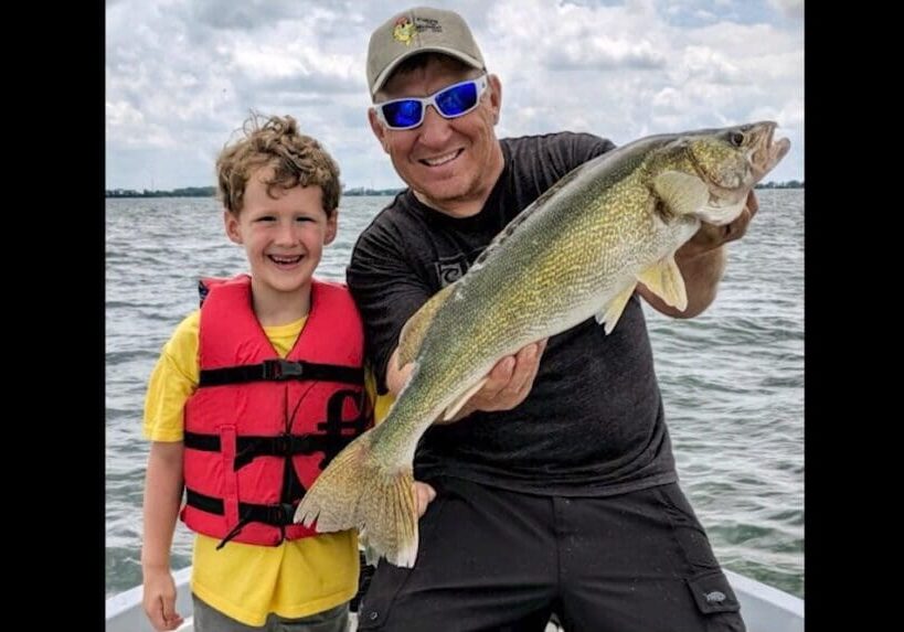 A smiling adult wearing sunglasses and a cap holds a large fish after a successful fishing trip, standing next to a smiling child in a red life jacket on a boat, with water and a cloudy sky in the background.