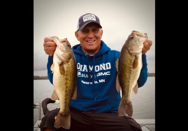 A smiling man, Mike Frisch, in a blue hoodie and cap holds up two large fish, one in each hand, while standing on a boat with water and an overcast sky in the background.