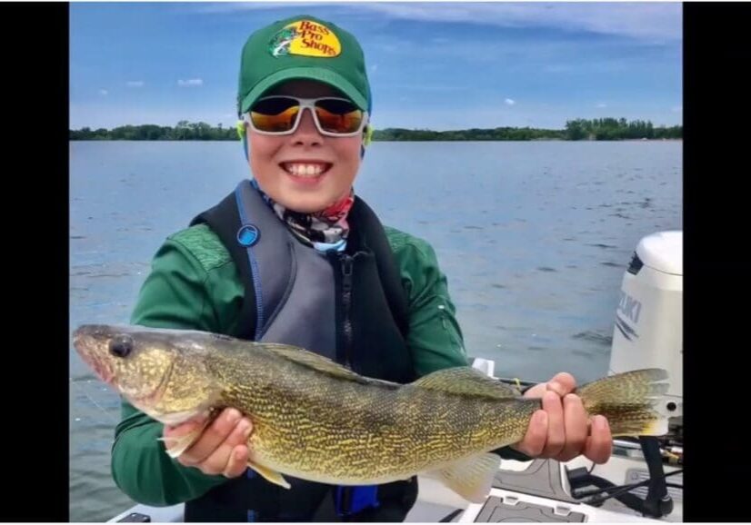 A smiling child wearing sunglasses and a green cap holds a large fish on a boat after a fun day of fishing, with a lake and trees in the background under a blue sky.