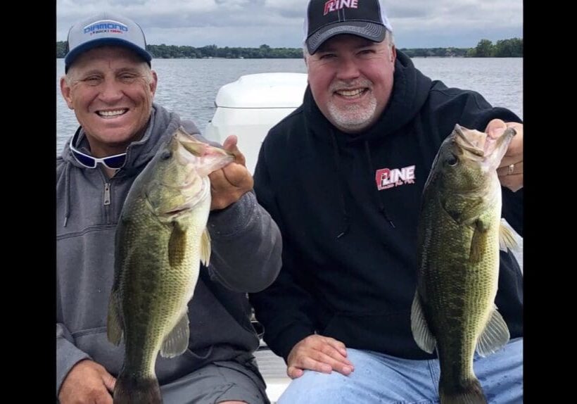 Two smiling men sit on a boat holding large bass fish, with a lake and cloudy sky in the background. Both wear caps and casual outdoor clothing, enjoying a successful fishing trip and appearing happy with their catch.