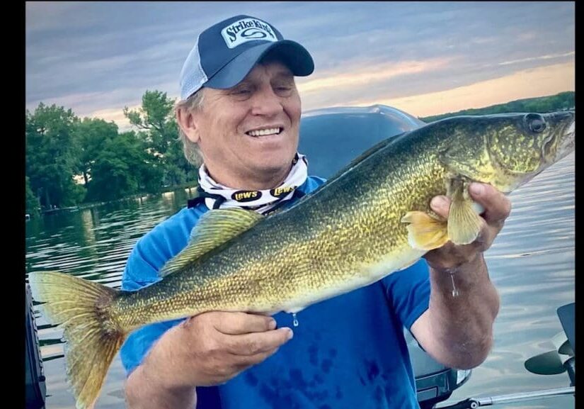 A smiling man in a blue shirt and cap holds a large, golden-brown fish, proudly showing off his fishing catch by the lake, with trees and a cloudy sky in the background.