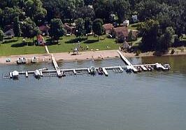 Aerial view of Midway Beach Resort with docks extending into the water, several boats docked, a sandy beach, grassy lawns, and houses surrounded by trees in the background.