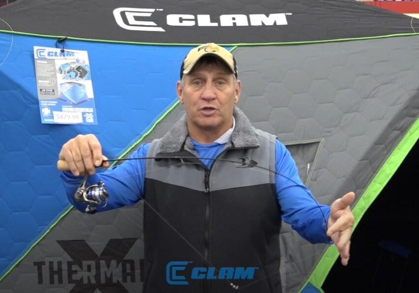 A man in a blue shirt and cap holds a fishing rod in front of a gray and blue CLAM ice fishing shelter with green trim. A product sign and the CLAM logo are visible on the shelter behind his fishing setup.