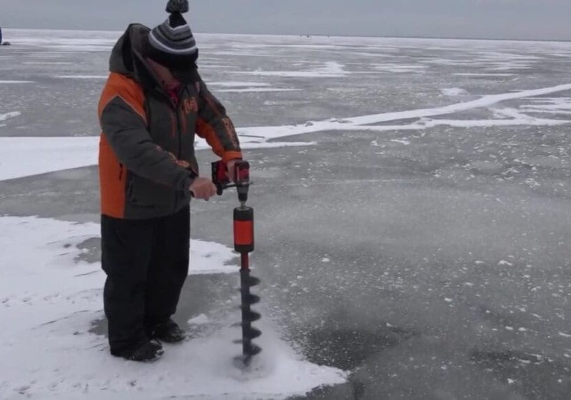 A person dressed in winter clothing uses an auger to drill a fishing hole in the ice on a frozen lake, with snow and ice covering the surface and a blue tent visible in the distance.