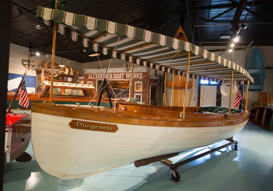 A vintage wooden boat named "Dungeness" with a striped canopy is displayed indoors at the Legacy of the Lakes Museum, with an American flag on the stern and an "Alexandria Boat Works" sign in the background.