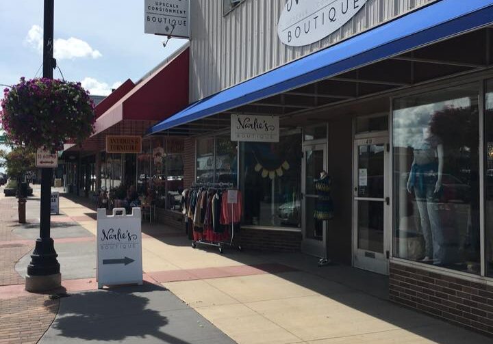 A sidewalk view of Narlie's Upscale Consignment Boutique, with a blue awning, clothing displays outside, a hanging flower basket, and nearby storefronts on a sunny day.