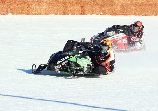 Two racers on snowmobiles speed around a bend on a snowy track, leaning into the turn as the spectacular tradition continues. The lead snowmobile is black and green, while the second is red and white. Both riders wear helmets and racing suits.