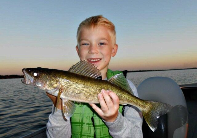 A smiling young boy in a green life jacket holds a freshly caught fish on a boat, embodying Fall Fishing Trends in Alexandria, with calm water and a sunset sky in the background.