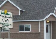 Northwoods Cafe is a brown building with white trim, featuring a peaked roof and visible windows and door. Out front, a sign reads "Northwoods Cafe, Breakfast & Lunches, 763-2900," welcoming guests inside.