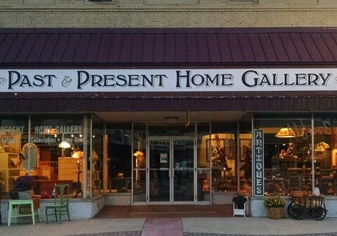 Front view of Past & Present Home Gallery, featuring large glass windows displaying antiques and home decor. A sign reading "ANTIQUES" and cozy lighting invite visitors inside.