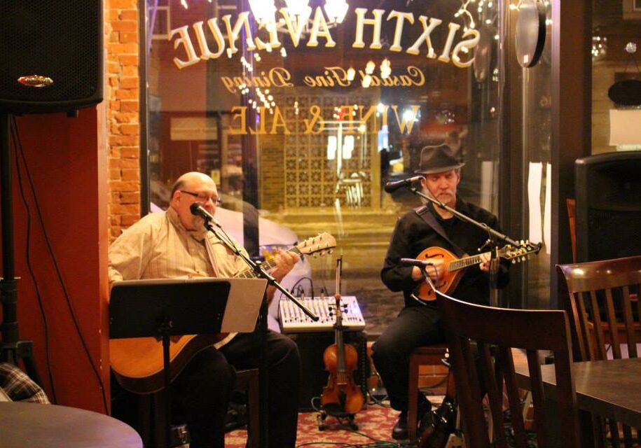 Two musicians perform live music in a cozy, dimly lit café in Alexandria, MN. One plays an acoustic guitar and sings, while the other strums a mandolin near the window of "Sixth Avenue Café and Wine Bar." Empty chairs sit in the foreground.