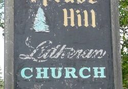 A weathered wooden sign reading "Spruce Hill Lutheran Church," featuring a small tree graphic, stands outdoors with Spruce Hill Lutheran Church nestled among the surrounding trees.