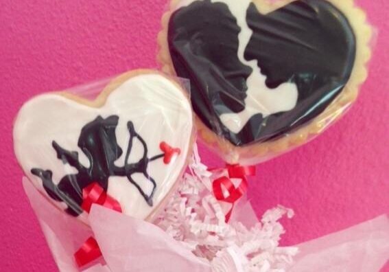 Two heart-shaped cookies on sticks, decorated with black and white icing—one featuring a couple’s silhouette, the other an abstract design with a red heart. Perfect for Valentine's Weekend in Alexandria, both are wrapped in plastic against a pink background.
