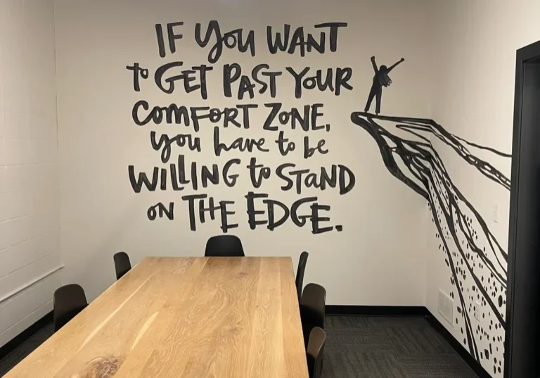 A modern conference room by The Edge Co features a wooden table, black chairs, and a mural reading, "If you want to get past your comfort zone, you have to be willing to stand on the edge," with art of a person on a cliff.