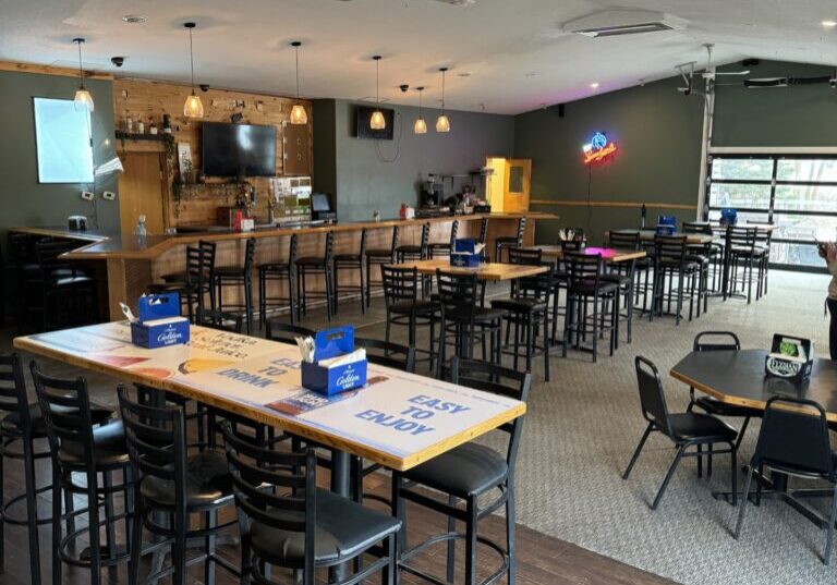Spacious bar and restaurant interior with high tables and chairs, a long bar counter, TV, neon signs, and large windows providing natural light. The Locals offers clean, modern Dining in one of the top Alexandria restaurants, warmly lit for a welcoming vibe.